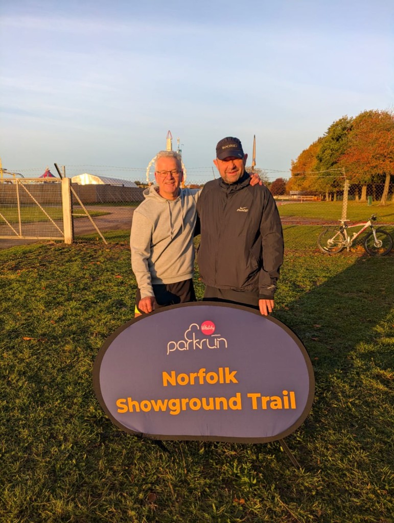 Two Norfolk Run Collective runners with the parkrun Norfolk Showground Trail sign